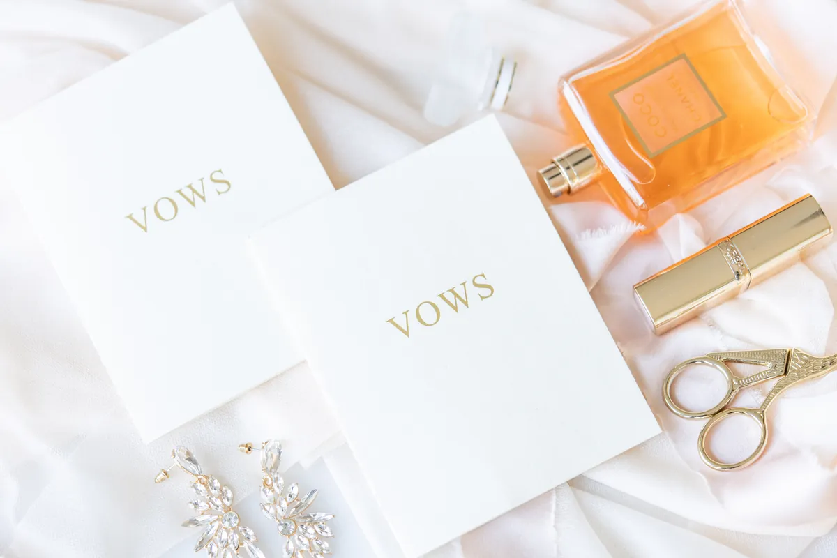 Two gold-foil “Vows” books styled with perfume, earrings, and gold details on white linen.